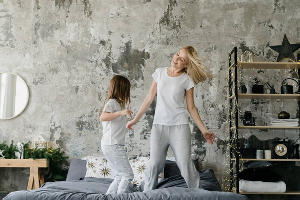 Ganztägige Ansicht der glücklichen Mutter, die zusammen mit ihrer Tochter auf dem Bett springt, während sie den Tag zusammen zu Hause verbringen. Frau und Mädchen im Pyjama amüsieren sich und tanzen morgens im Schlafzimmer - Foto, Bild