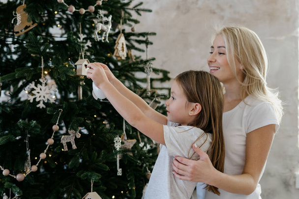Winter holidays concept. Profile view of little girl holding new year toy and decorate christmas tree, preparation for new year and christmas while happy mother support and standing near daughter - 写真・画像