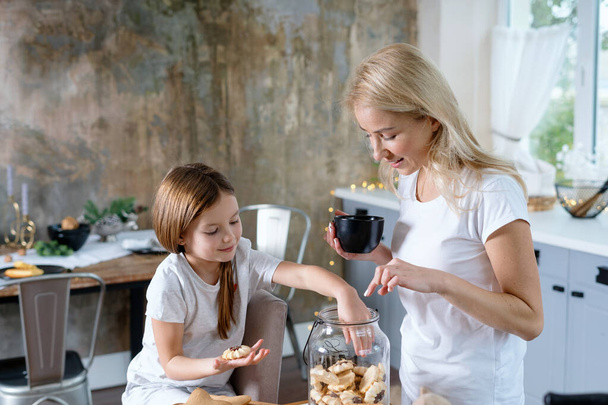 Loving mother holding cup with beverage in hand, spending morning at kitchen together with little daughter. Smiling girl taking cookie from glass jar before breakfast - Valokuva, kuva