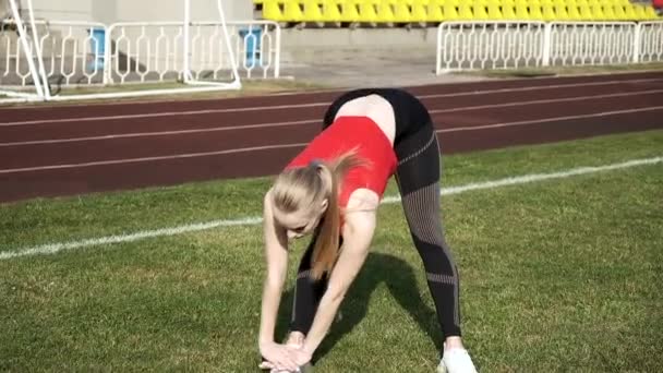 młoda kobieta w sportowej odzieży rozciągającej ciało na tle stadionu sportowego - Materiał filmowy, wideo
