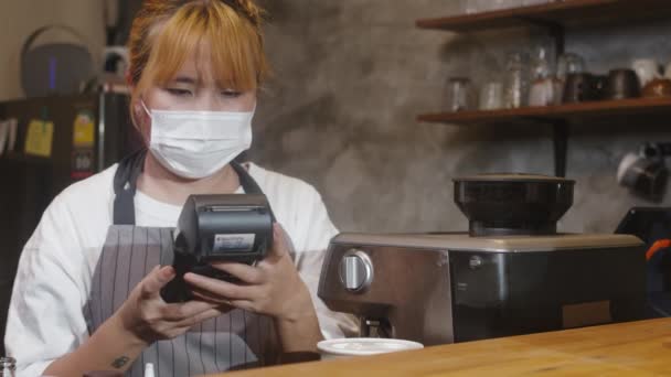 Young Asia female barista wear face mask serving take away hot coffee paper cup to consumer and using credit card for payment at cafe. Lifestyle new normal after corona virus and social distancing. - Footage, Video