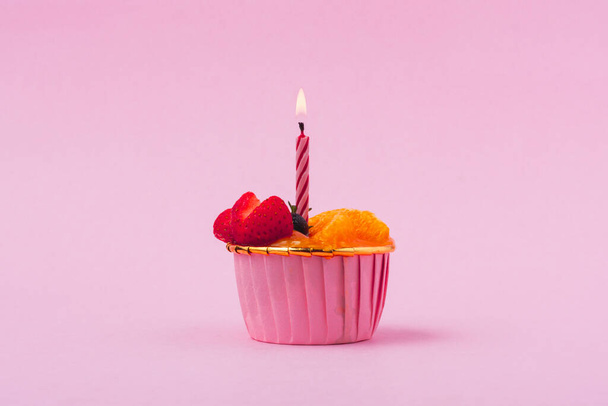 Мини бумажная чашка фруктового или апельсинового бисквита с горящей свечой на розовом фоне для праздничной выпечки, питания и питания - Фото, изображение