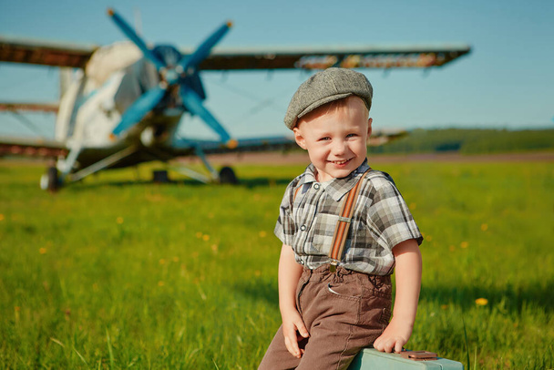Little boy, happy kid with a small suitcase on the background of planes on the airfield. The child dreams of travelling and flying on an airplane. His future profession is pilot - Foto, afbeelding