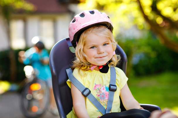 Portrait of little toddler girl with security helmet on the head sitting in bike seat of parents. Boy on bicycle on background. Safe and child protection concept. Family and weekend activity trip. - Photo, Image
