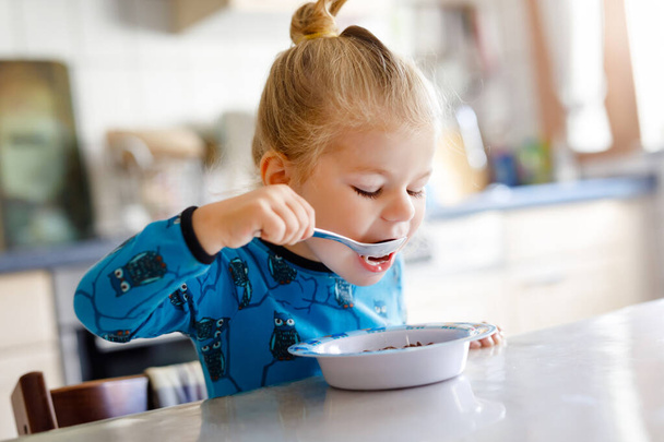 Küçük, güzel bir kız kahvaltıda sağlıklı mısır gevreği ve süt yiyor. Renkli giysiler içinde şirin bir bebek mutfakta oturup yulaf ve mısır gevreği hazırlarken eğleniyor. Evde kapalı. - Fotoğraf, Görsel