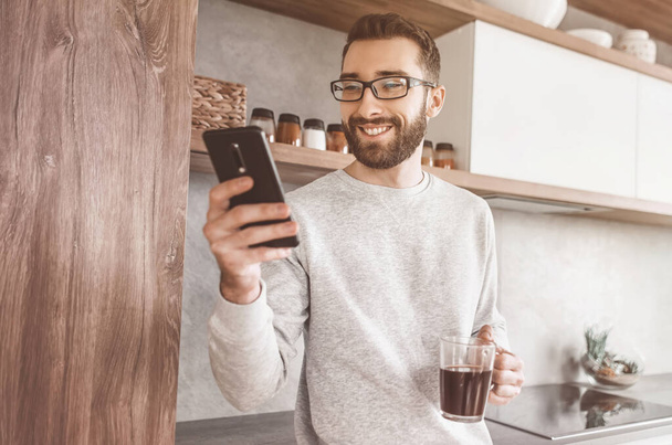 smiling man with smartphone and Cup of coffee standing in his kitchen - Fotoğraf, Görsel
