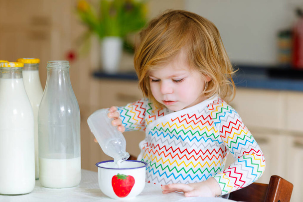 愛らしい幼児の女の子は朝食のために牛乳を飲んでいます。かわいい赤ちゃんの娘にボトルがたくさんあります。健康的な子供はカルシウム源としてミルクを持っています。午前中に家や保育園で子供. - 写真・画像
