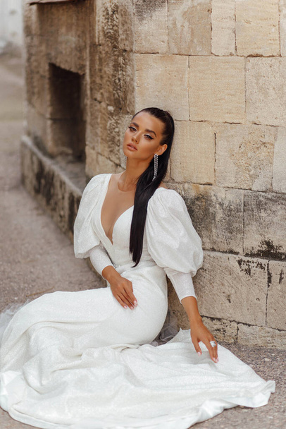 fashion outdoor photo of beautiful woman with dark hair in luxurious wedding dress and accessories posing in the summer street - Fotó, kép