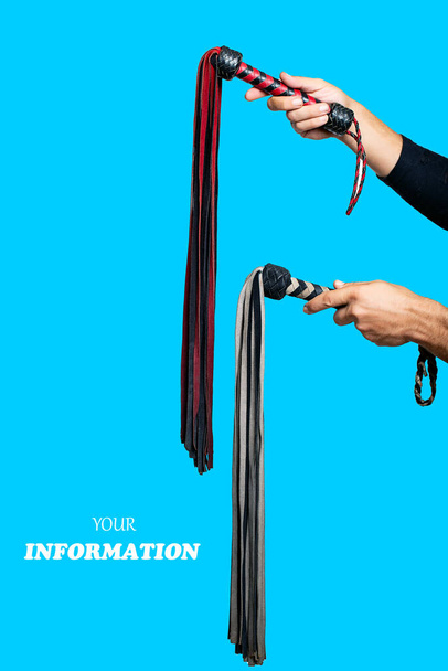 Two leather whips in male hand. Man's hand is holding bdsm lash isolated on blue background. Accessories for adult sexual games. Toys for BDSM, flogging, sexual fetish and spanking devices - Photo, Image