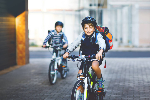 Two school kid boys in safety helmet riding with bike in the city with backpacks. Happy children in colorful clothes biking on bicycles on way to school. Safe way for kids outdoors to school - 写真・画像