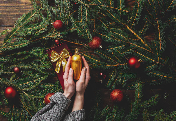 Vrouwelijke handen houden parfum fles naast kerstboom takken en kerstballen rond - Foto, afbeelding