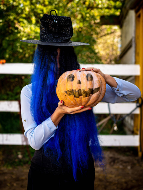 Egy lány, akinek boszi jelmezben van a haja, egy festett arcú tököt és egy boszorkány sapkát tart a kezében. - Fotó, kép