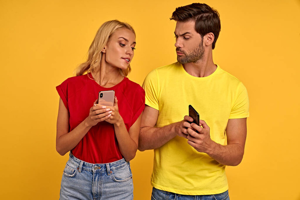 Glimlachend nieuwsgierig jong koppel vrienden jongen meisje in gekleurde t-shirts poseren geïsoleerd op gele achtergrond studio. Mensen levensstijl concept. Een jaloers meisje kijkt in de telefoon van haar vriend. - Foto, afbeelding