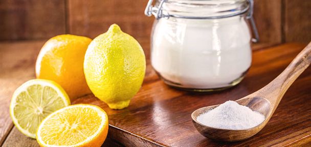 Glass pot with baking soda and wooden measuring spoon, with fruits in the background, lemon and orange, chemical compound used in industry and cooking, as an antacid or as yeast - Photo, Image