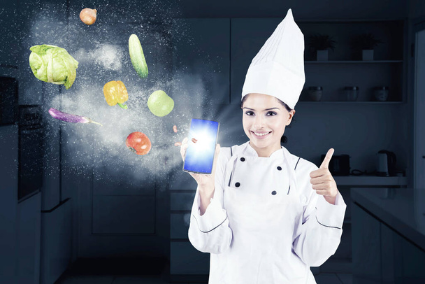 Chef mujer mostrando el pulgar hacia arriba mientras muestra un teléfono celular con ingredientes voladores de alimentos y de pie en la cocina - Foto, imagen