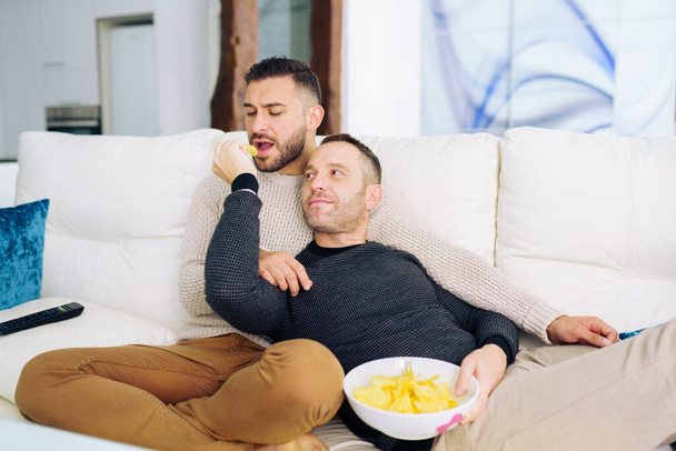 Gay pár sedí doma na gauči a dívá se na něco v televizi a dává si svačinu. - Fotografie, Obrázek