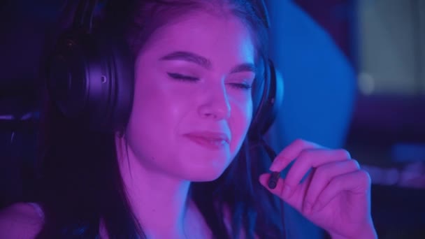 Νεαρή όμορφη γυναίκα κάθεται στο νέον gaming club - φορώντας μεγάλα ακουστικά και μιλώντας στο μικρόφωνο - Πλάνα, βίντεο