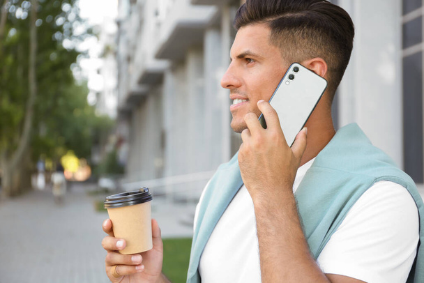 Man with cup of coffee talking on modern mobile phone outdoors - Foto, afbeelding