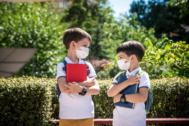 dva chlapci, školáci v černých školních uniformách s batohy v lékařské masce jít do školy samostatně, držet se za ruce, přátelství koncept, studijní koncept během coronavirus, zpět do školy. - Fotografie, Obrázek