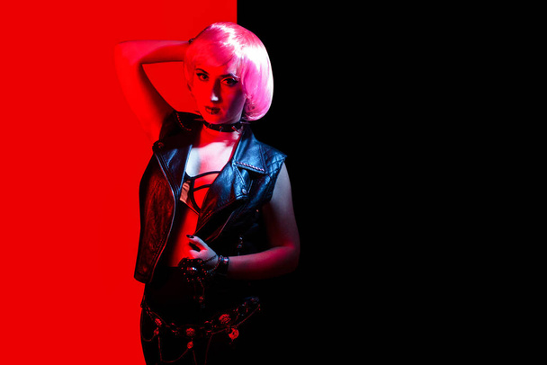 Young beautiful rocker girl with pink hair and lip piercing wearing leather vest and spiked collar on red and black background. - 写真・画像