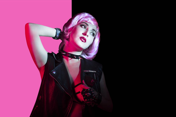 Beautiful young rocker woman with pink hair and lip piercing wearing leather vest and spiked collar on pink and black background. - Photo, Image