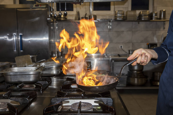 Flame in the pan. Professional Chef makes flambe for food in the restaurant kitchen. Chef cooking with open fire pan on a stove.  - Photo, Image
