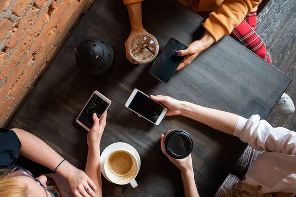 Верхний вид женских рук со смартфонами и чашками кофе на столе в кафе. Три девушки пьют кофе. - Фото, изображение