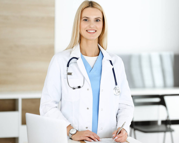 Woman-doctor at work in clinic happy of her profession. Blond female physician controls medication history record and medical exam results. Medicine concept - Zdjęcie, obraz