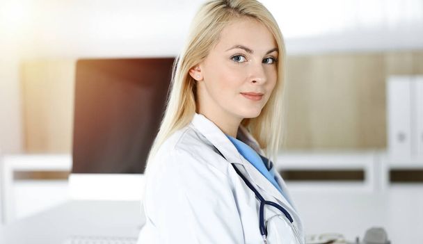 Woman-doctor at work while sitting at the desk in sunny clinic. Blonde cheerful physician ready to help - Foto, imagen