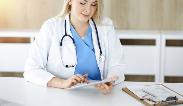 Woman-doctor at work while sitting at the desk in sunny clinic. Blonde cheerful physician filling up medication history record form - 写真・画像