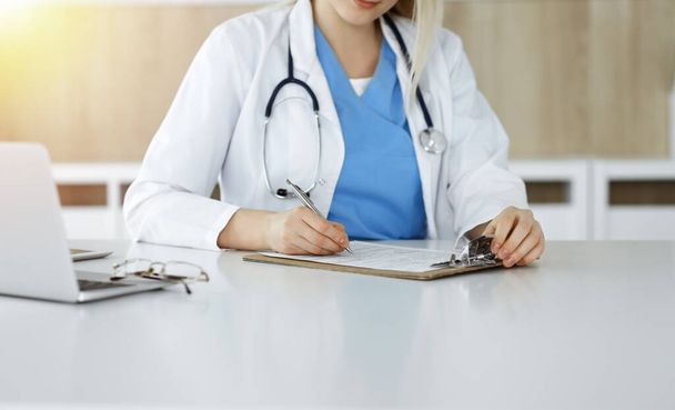 Unknown woman-doctor at work filling up medication history record form in sunny clinic, close-up - Foto, Imagen