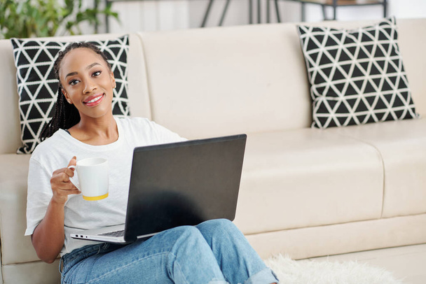 Positieve mooie jonge zwarte vrouw met grote mok drinken van koffie bij het programmeren op laptop thuis - Foto, afbeelding