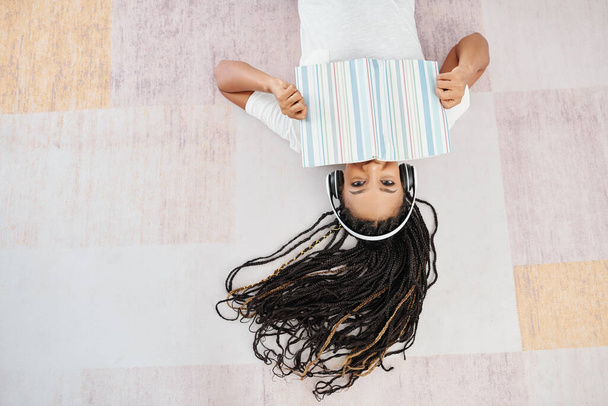Happy pretty young Black woman with dreadlocks lying on the floor and covering mouth with opened book - Fotó, kép