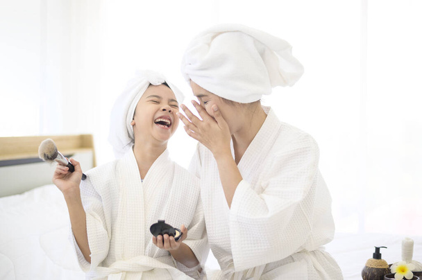 A happy beautiful mom and daughter in white bathrobe applying natural Make-Up with cosmetic powder brush, Family and Beauty Concept.  - Фото, изображение