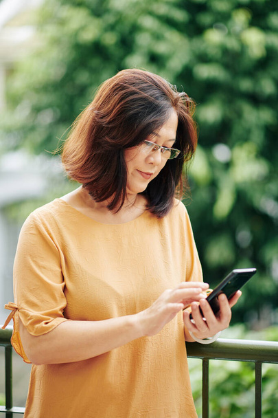 Зріла в "єтнамська жінка в окулярах перевіряє текстові повідомлення у смартфоні. - Фото, зображення
