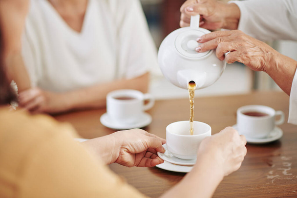 Hände einer Seniorin gießen ihren Gästen bei Party heißen Tee in Tassen ein - Foto, Bild