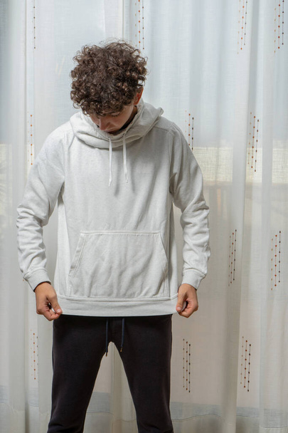 Attractive young man with curly hair wearing white sweatshirt posing on white curtains background. Fashion concept - Φωτογραφία, εικόνα