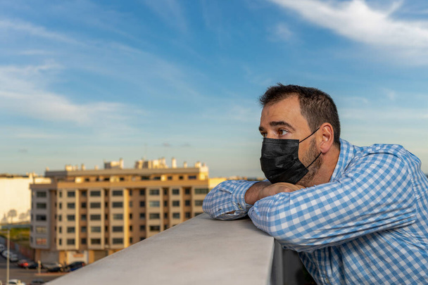 Young man on the balcony of the house during the covid-19 outbreak. 40 year old man with mask, worried and tired face looking at the street thoughtful and depressed in quarantine. Isolation concept - Photo, Image