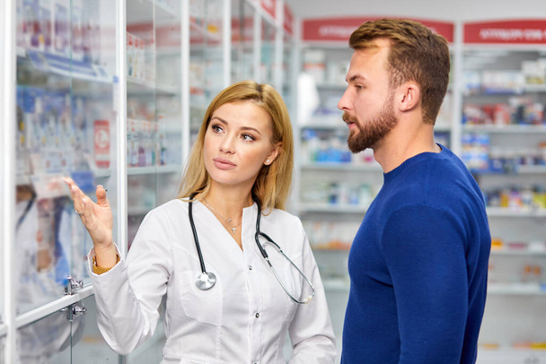 young female pharmacist helping male customer choosing prescription drugs from the shelf - Foto, Bild