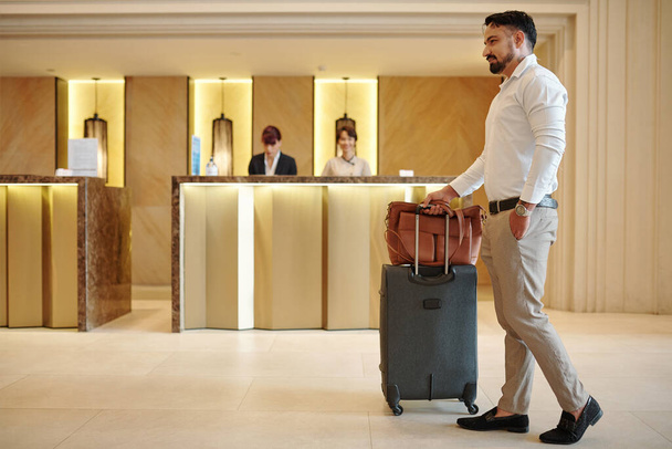 Jóképű, elegáns férfi aktatáskával és bőrönddel belép egy drága hotel halljába. - Fotó, kép