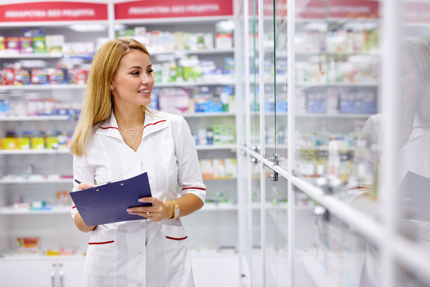 blonde druggist woman in uniform checking the assortment in pharmacy - Foto, imagen
