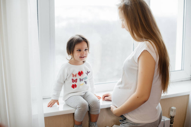 A pregnant mother is standing near the window with her little daughter - Valokuva, kuva