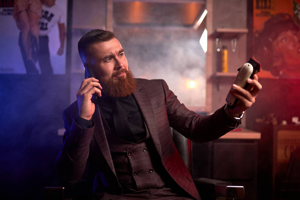 young male in suit, barber man hold trimmer in hands and talk on phone - Foto, imagen