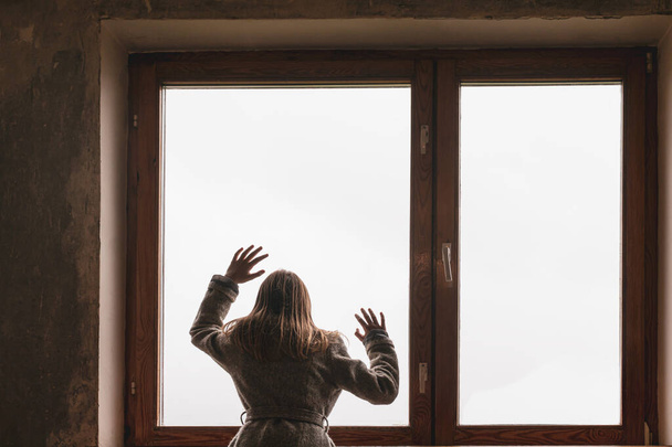 Έγχρωμη εικόνα ενός νεαρού κοριτσιού που στέκεται κοντά σε ένα παλιό παράθυρο και είναι ντυμένο με ζεστά ρούχα - Φωτογραφία, εικόνα