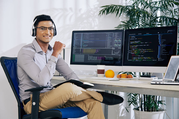 Ritratto di sorridente fiducioso programmatore indiano seduto alla scrivania del suo ufficio con codice di programmazione sugli schermi del computer - Foto, immagini