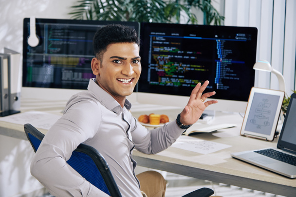 Desarrollador de software indio emocionado feliz sentado en el escritorio de la oficina, dando la vuelta y saludando a la cámara - Foto, Imagen