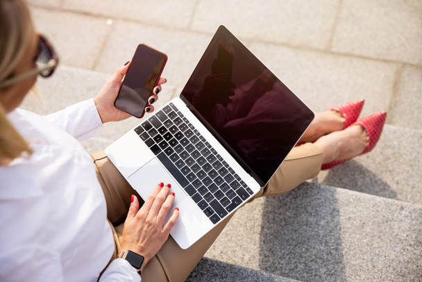 Жінка використовує ноутбук і мобільний телефон, сидячи надворі
 - Фото, зображення