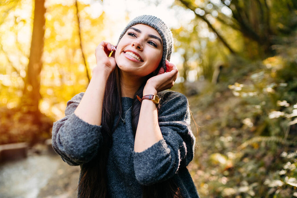 Very beautiful and happy woman in the autumn season in a forest with lots of light with the leaves of the orange and yellow trees. The woman is wearing warm clothes and a gray wool cap - Фото, изображение