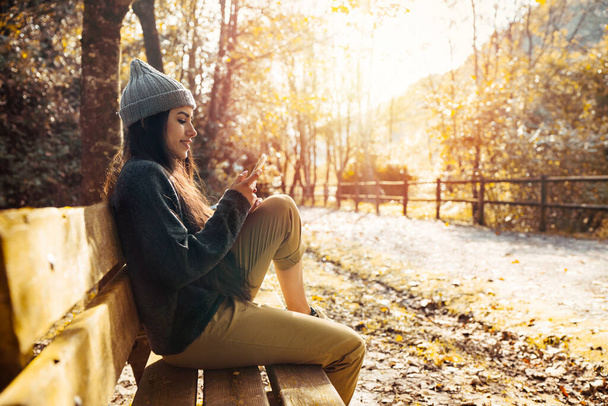 Bardzo piękna i bardzo szczęśliwa kobieta w sezonie jesiennym siedzi na ławce leśnej drogi z liśćmi drzew pomarańczowych i żółtych.Kobieta rozmawia przez telefon komórkowy w ciepłych ubraniach - Zdjęcie, obraz