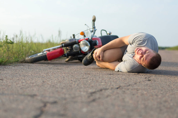Um homem segura no joelho, deitado no asfalto perto de sua motocicleta, o tema dos acidentes rodoviários - Foto, Imagem